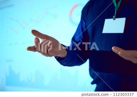Professional in blue sweater gesturing with hands in front of screen with presentation with charts and data. 112303337