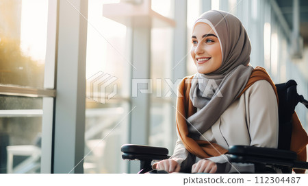 Happy, smiling young Muslim girl student in a wheelchair in the foyer of an educational institution. Happy, smiling young Muslim girl student in a wheelchair in the foyer of an educational institution. 112304487