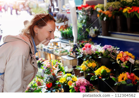 Young adult joyful woman kneels to choose buy colorful potted flowers at outdoor market stall. Sale of blooming plants in pots at botanical street German store on sunny spring day Young adult joyful woman kneels to choose buy colorful potted flowers at outdoor market stall. Sale of blooming plants in pots at botanical street German store on sunny spring day 112304562
