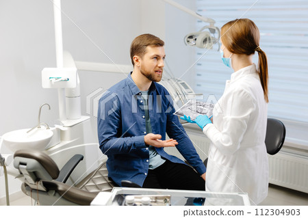 Young attractive man visiting dentist, sitting in dental chair at modern light clinic. Young woman dentist holding x ray image. 112304903