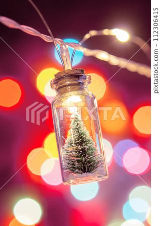 Merry Christmas and happy new year. Hanging small Christmas tree in glass jar on pine branches Christmas tree garland and ornaments over abstract bokeh 112306415