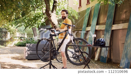 Youthful man in home yard carrying his toolbox and preparing for bike summer maintenance. Side-view portrait of caucasian male holding different specialized equipments to repair bicycle. 112310696