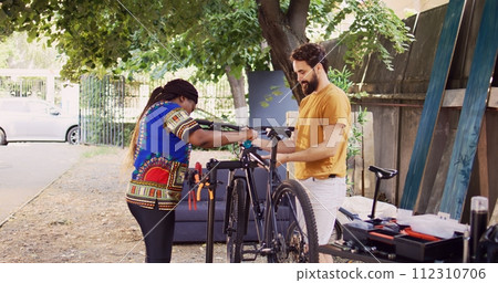 Sports-loving multiethnic couple fastening bike body for examination and maintenance. African american girlfriend assisting boyfriend by clutching modern bicycle to repair-stand in home yard. 112310706