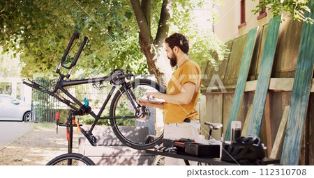 Male cyclist holding personal computer and inspects bike pedal and crank arm for damages in home yard. Active caucasian man enjoys browsing internet, fixing and maintaining modern bicycle. Male cyclist holding personal computer and inspects bike pedal and crank arm for damages in home yard. Active caucasian man enjoys browsing internet, fixing and maintaining modern bicycle. 112310708