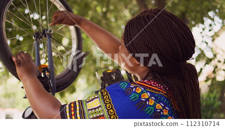 Healthy black woman mending her own bicycle using specialized equipment from toolkit in yard. Dolly zoom-in shot. Female african american cyclist repairing front bike wheel outdoors. 112310714