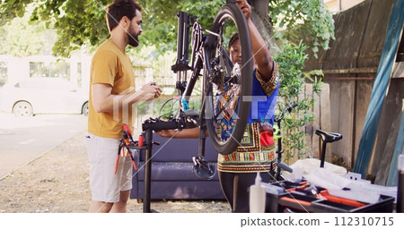 Committed interracial couple outside servicing bicycle components. Healthy active black woman and caucasian man inspecting and maintaining bike tires and chains for leisure cycling. Pull-back shot. 112310715