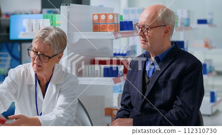 Pharmacist in drugstore with senescent patient after finishing annual examination, suggesting him cardivascular pills to treat heart disease symptoms. Aged man receiving treatment from specialist Pharmacist in drugstore with senescent patient after finishing annual examination, suggesting him cardivascular pills to treat heart disease symptoms. Aged man receiving treatment from specialist 112310730