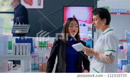 Proficient healthcare practitioner in apothecary helping customer with medical suggestions. Client in dispensary wishing to purchase supplements and vitamins, asking druggist for guidance 112310735