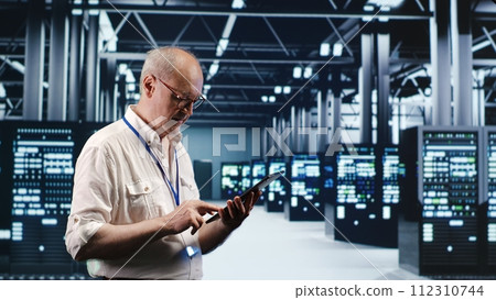 Senescent professional navigating network of server rigs in industrial nexus. Employee with tablet doing analysis and optimizations in high tech data center, preventing system perils 112310744