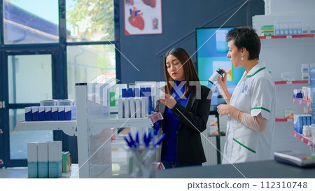 Shopper in chemist store asking pharmacist for medical expertise while she arranges merchandise on shelves. Licensed druggist helping customer choose best supplements for her ailment 112310748
