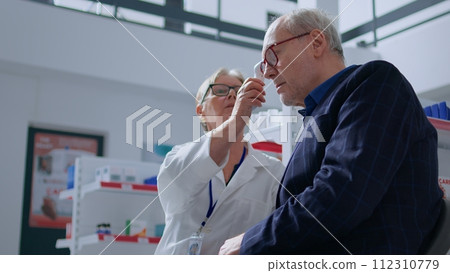 Elderly pharmacist in drugstore using electronic thermometer on senior man, checking temperature. Old patient and healthcare specialist during yearly examination, monitoring flu symptoms such as fever Elderly pharmacist in drugstore using electronic thermometer on senior man, checking temperature. Old patient and healthcare specialist during yearly examination, monitoring flu symptoms such as fever 112310779