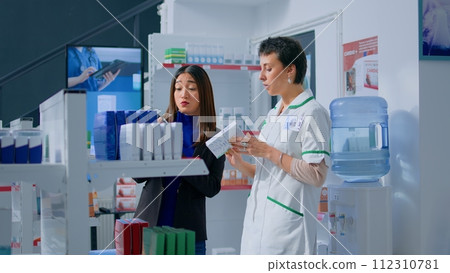 Shopper in chemist shop asking pharmacy employee about product information, trying to find right items on prescription list. Adept druggist helping customer choose fitting sickness medication 112310781