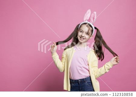 Young child acting joyful and happy about easter celebration, posing with confidence against pink background. Small toddler wearing bunny ears and pigtails, feeling positive and excited. 112310858