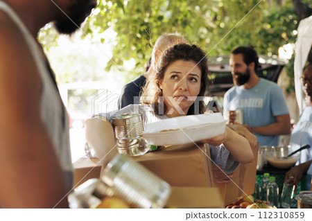Portrait shot of poor and needy caucasian woman carrying her donation box and canned goods from food bank. Image showing the underprivileged and homeless people receiving support from charity group. Portrait shot of poor and needy caucasian woman carrying her donation box and canned goods from food bank. Image showing the underprivileged and homeless people receiving support from charity group. 112310859