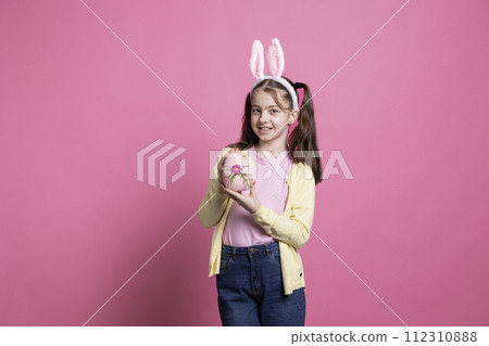 Small lovely child with bunny ears holding a painted pink egg in front of camera, presenting her handmade easter decoration over colorful background. Young adorable girl shows traditional ornaments. Small lovely child with bunny ears holding a painted pink egg in front of camera, presenting her handmade easter decoration over colorful background. Young adorable girl shows traditional ornaments. 112310888