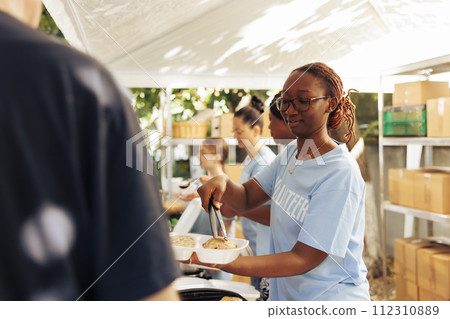African american woman serving free food to the poor, needy individual at an outdoor food bank. Female volunteers feeding and providing support to the hungry and less privileged at a homeless shelter. African american woman serving free food to the poor, needy individual at an outdoor food bank. Female volunteers feeding and providing support to the hungry and less privileged at a homeless shelter. 112310889