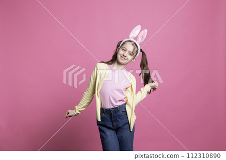 Cheery little girl dancing and having fun in front of camera, feeling happy and excited about easter celebration. Confident small toddler showing dance moves while she wears bunny ears. 112310890