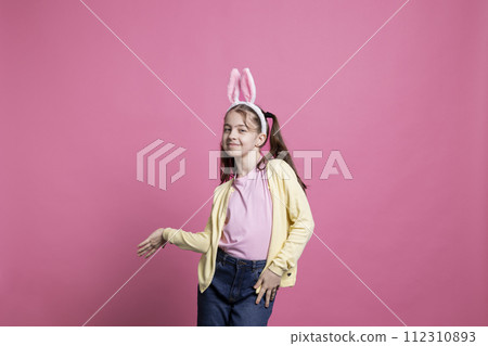 Joyful toddler with pigtails and bunny ears dancing on camera, showing cute dance moves and fooling around in studio. Sweet young girl feeling excited about easter celebration event. 112310893