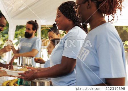 Voluntary multiethnic people distributes needed products and free meals to the hungry, demonstrating charity in aiding the less privileged. Non-profit outdoor food bank serving hot food to the needy. 112310896
