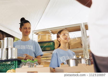 Caucasian mother and daughter volunteering at food drive, providing free food to the poor and needy. Female charity workers helping and giving essential items to homeless people and less fortunate. 112310897