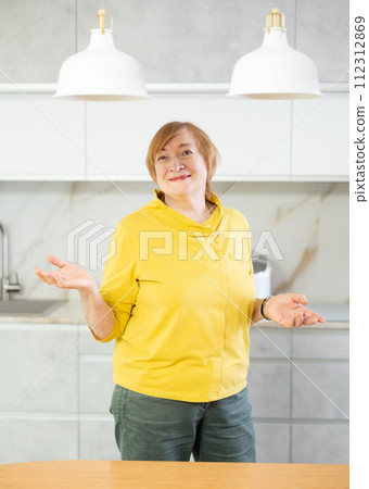 Smiling senior woman in casual clothes standing in home kitchen with open arms 112312869