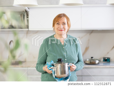 Elderly woman stands in modern home kitchen and holds pan with dishcloth in hands 112312871
