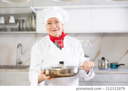 Elderly woman in chef hat and white cook uniform stands near kitchen table and holds frying pan Elderly woman in chef hat and white cook uniform stands near kitchen table and holds frying pan 112312879