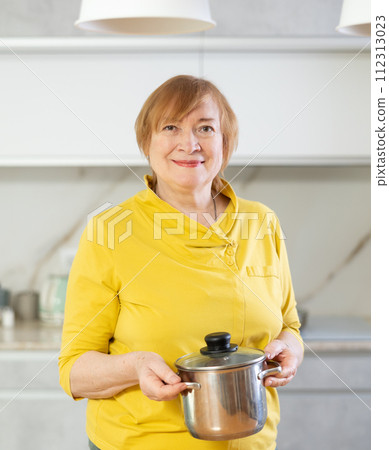 Elderly woman in casual clothes stands in front of table and holds stainless pan with lunch 112313023