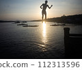Silhouette of a man in shorts jumping into the water at a seaside pier. 112313596