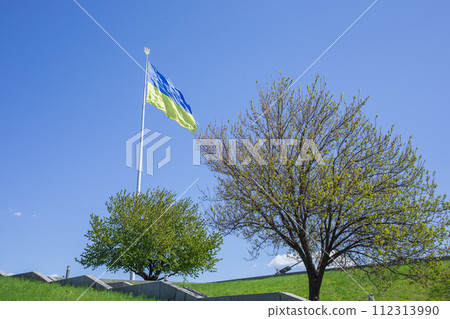 [Ukraine] Capital Kiv, huge Ukrainian flag flutters against the blue sky, fresh green grass and street trees 112313990