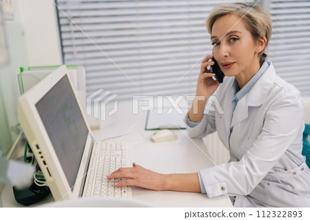 Portrait of middle-aged Caucasian female doctor in white coat talking on smartphone in hospital clinic sitting at desk with laptop, looking at camera. Woman medical worker physician have conversation Portrait of middle-aged Caucasian female doctor in white coat talking on smartphone in hospital clinic sitting at desk with laptop, looking at camera. Woman medical worker physician have conversation 112322893
