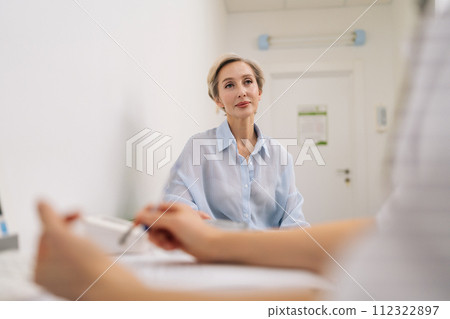Portrait of middle-aged female patient telling to unrecognizable doctor about illness in medical facility. Rear view of physician explaining treatment, recommending therapy, giving medical help. 112322897