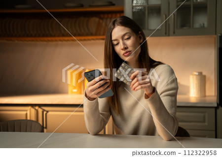 Young Woman Checking Her Phone While Holding Medication in a Kitchen Setting at Dusk Young Woman Checking Her Phone While Holding Medication in a Kitchen Setting at Dusk 112322958