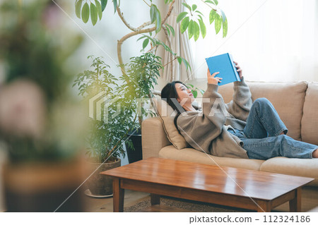 A woman reading while sitting on a sofa surrounded by greenery 112324186
