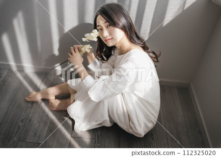 A woman in a white dress sitting on the floor by the window and holding flowers A woman in a white dress sitting on the floor by the window and holding flowers 112325020