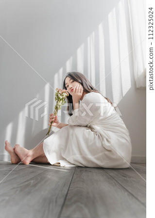 A woman in a white dress sitting on the floor by the window and holding flowers 112325024