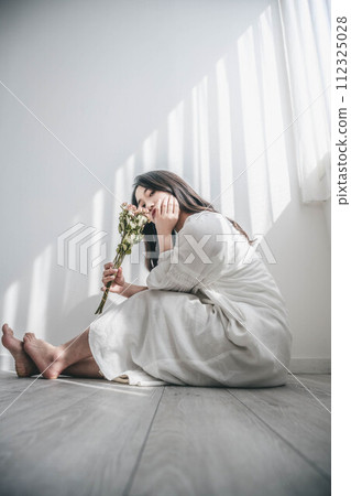A woman in a white dress sitting on the floor by the window and holding flowers 112325028