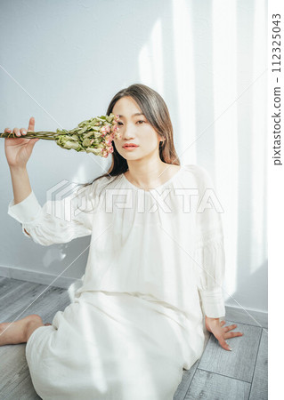 A woman in a white dress sitting on the floor by the window and holding flowers 112325043