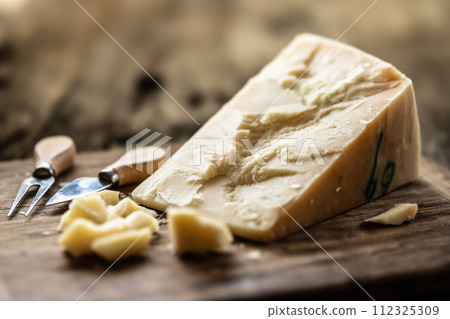 A block and pieces of Parmesan cheese on a cutting board along with a chisel and a skewer 112325309
