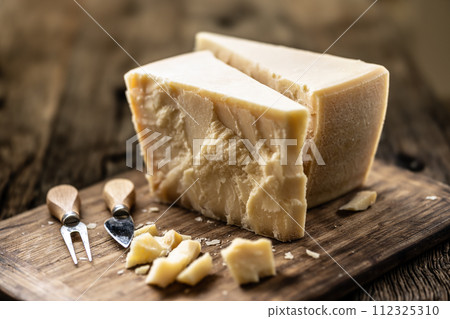 A block and pieces of Parmesan cheese on a cutting board along with a chisel and a skewer 112325310