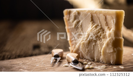 A block and pieces of Parmesan cheese on a cutting board along with a chisel and a skewer 112325312