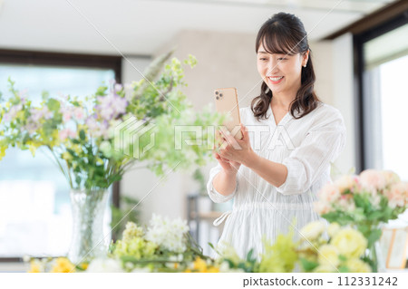 一位中年婦女在客廳用智能手機拍攝她愛好的花藝 112331242