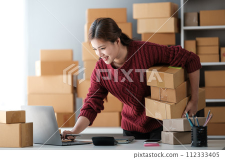 Startup SME small business entrepreneur of freelance Asian woman wearing apron using laptop and box to receive and review orders online to prepare to pack sell to customers, online sme business ideas. 112333405