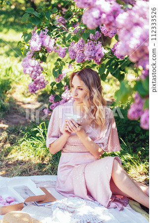 Serene Moment Under Lavender Lilac Blooms in Springtime. woman relaxes with a cup under blooming lilac bushes, enjoying a peaceful spring day. 112333526