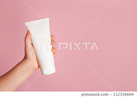 Child little hand holding white cream tube on pink background. Daily children beauty product. Closeup. Empty place for text or logo 112336604