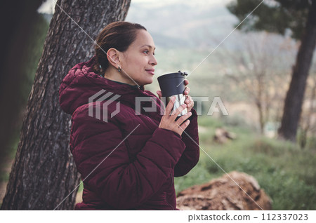 Young woman holding a thermos mug, enjoying spring nature in the forest, smiling and dreamily looking into the distance Young woman holding a thermos mug, enjoying spring nature in the forest, smiling and dreamily looking into the distance 112337023