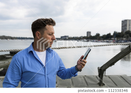 Man holding smartphone in his hand beside the river at port. Nautical and marine concept Man holding smartphone in his hand beside the river at port. Nautical and marine concept 112338346