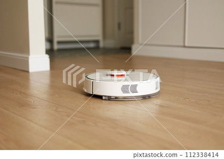 Modern wireless autonomous robotic vacuum cleaner cleans living room. Smart self-propelled white cleaning robot washes wooden floor. Household appliances with remote control. Modern wireless autonomous robotic vacuum cleaner cleans living room. Smart self-propelled white cleaning robot washes wooden floor. Household appliances with remote control. 112338414