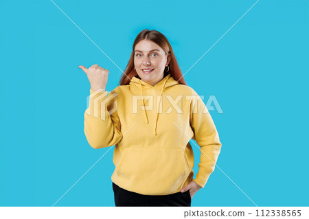Photo of red haired woman smiling showing thumb empty space recommending isolated blue color background, studio portrait Photo of red haired woman smiling showing thumb empty space recommending isolated blue color background, studio portrait 112338565