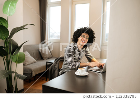 Smiling young man in striped shirt sitting at the table and having coffee Smiling young man in striped shirt sitting at the table and having coffee 112338793